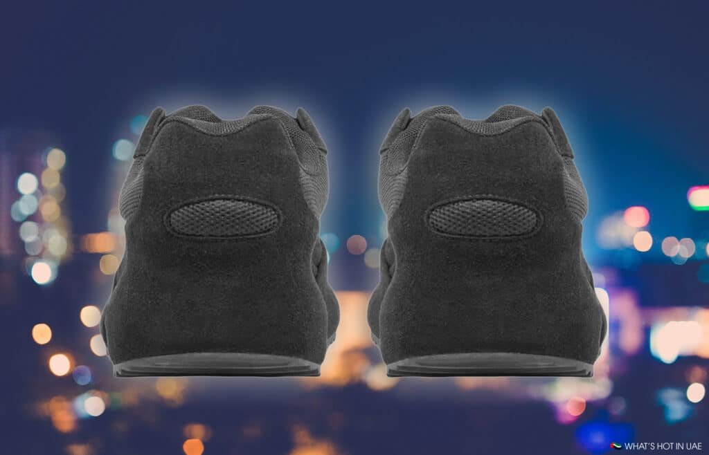 A pair of black sneakers shown from the back with a blurred city skyline at night in the background.