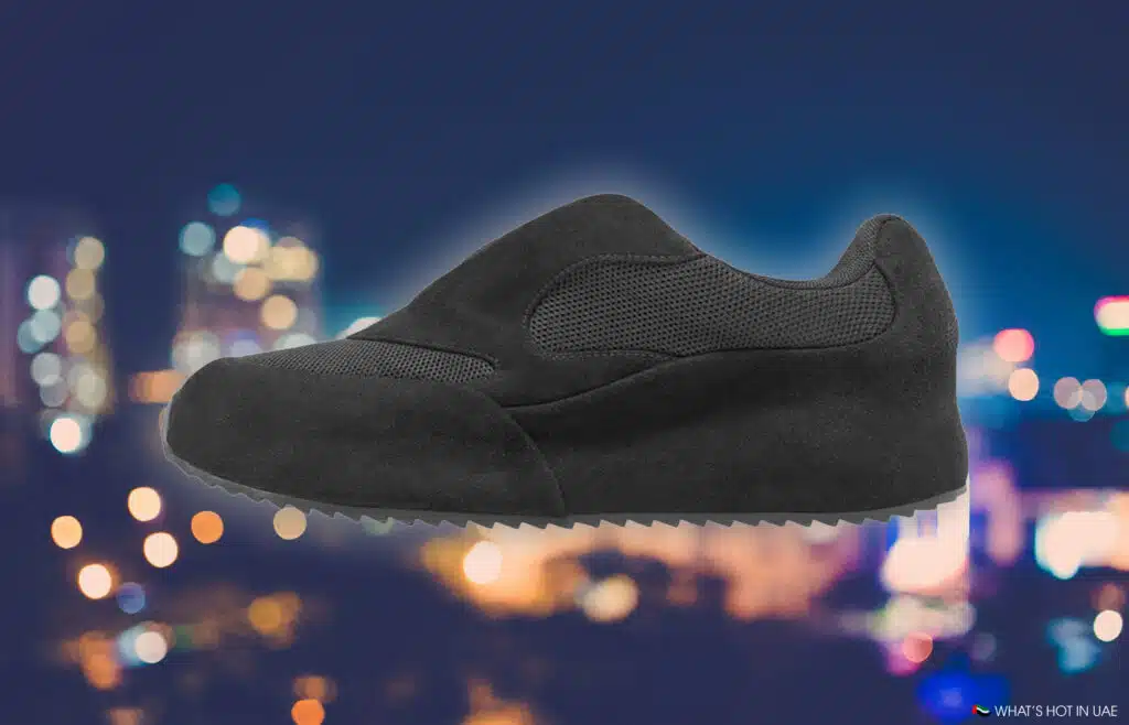A black sneaker with a suede and mesh design is shown against a blurred cityscape background.