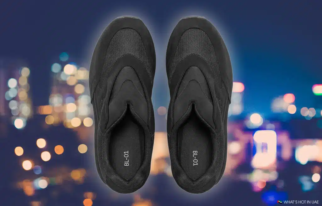 A pair of black YEEZY BL-01 slip-on sneakers against a blurred cityscape background.