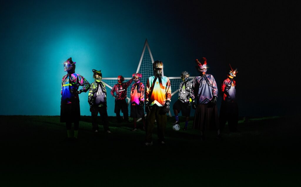 A group of individuals wearing futuristic and colorful outfits with helmets, standing in front of a soccer goal at night, illuminated against a dark blue sky.