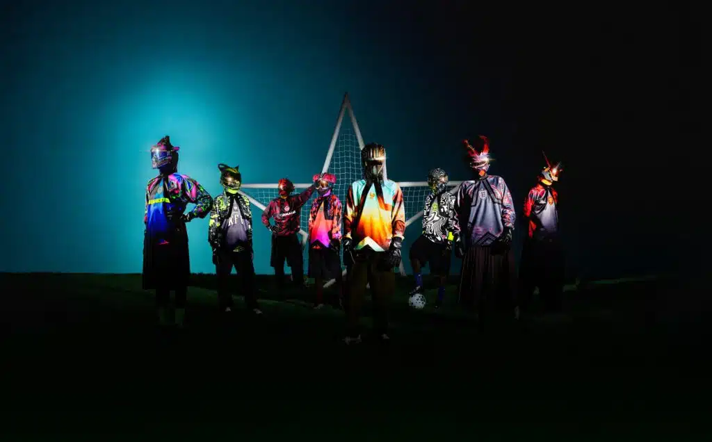 A group of individuals wearing futuristic and colorful outfits with helmets, standing in front of a soccer goal at night, illuminated against a dark blue sky.