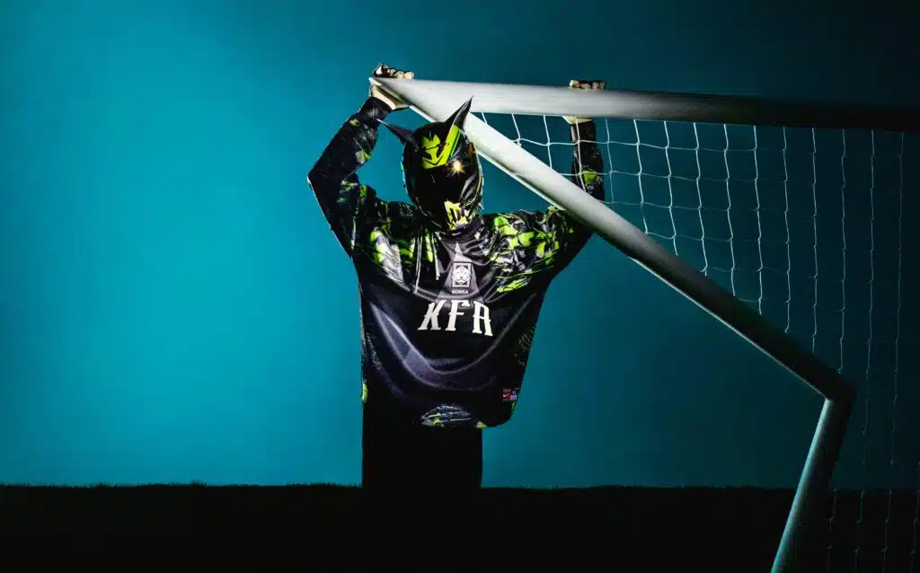 A person wearing a futuristic helmet and a patterned jersey with the letters 'KFR' stands dramatically in front of a soccer goal frame against a blue background.