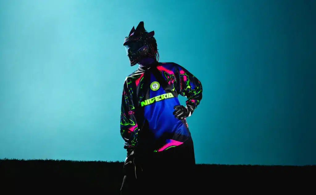 Nike Hollywood Keepers: A person wearing a colorful Nigeria-themed outfit with neon green and pink accents stands against a blue background. The outfit includes a stylized mask or helmet covering their head, contributing to a futuristic or fantasy appearance.