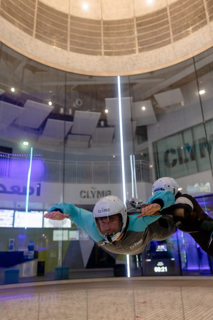 Chris Hemsworth in flight suits and helmets are indoor skydiving in a vertical wind tunnel.