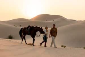 A man and woman walk alongside a horse in a desert with sand dunes at sunset.