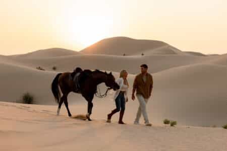 A man and woman walk alongside a horse in a desert with sand dunes at sunset.