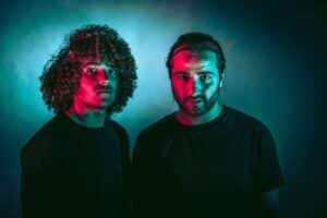 Two people in dark shirts are lit with dramatic red and blue lighting, creating a striking contrast against the dark background.