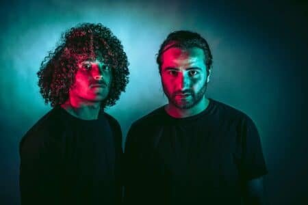Two people in dark shirts are lit with dramatic red and blue lighting, creating a striking contrast against the dark background.