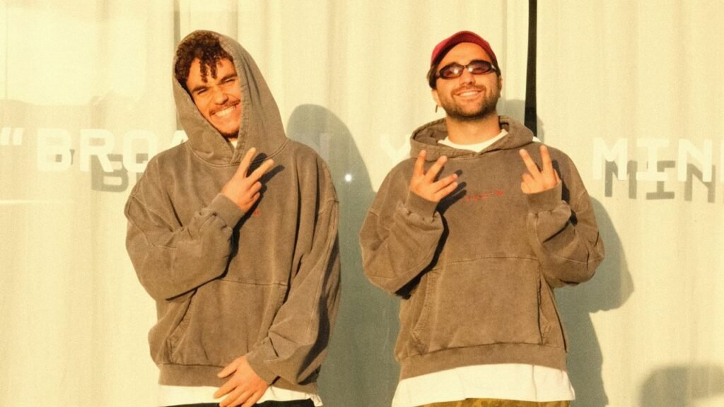 Two people wearing hoodies and making peace signs, standing against a wall with text partially visible in the background.