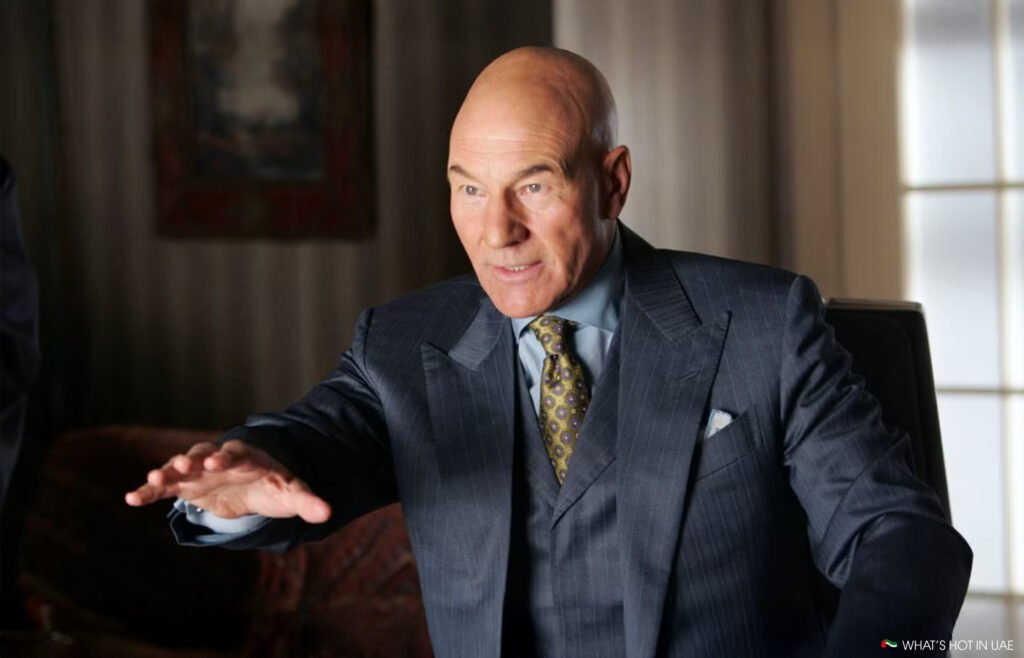 A bald man in a suit gestures with one hand, sitting in an office setting.