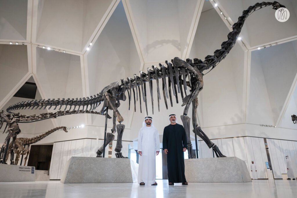 Two people in traditional Middle Eastern attire stand in front of a large dinosaur skeleton in Abu Dhabi Natural History Museum.