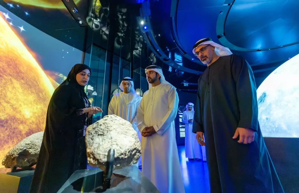 A group of people in traditional Middle Eastern attire observes a large rock exhibit in a modern, blue-lit space.