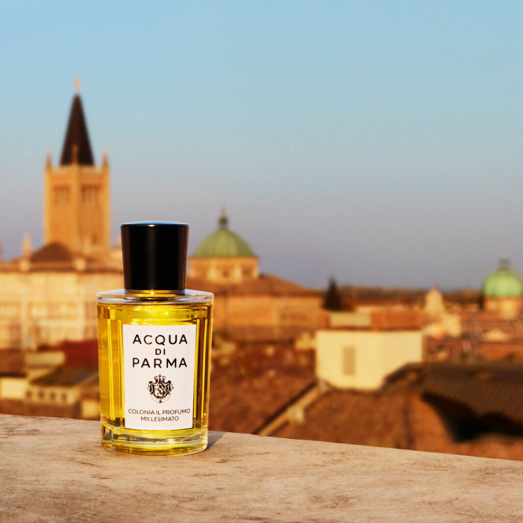 A bottle of Acqua di Parma perfume sits on a ledge, with an Italian cityscape featuring historical buildings in the background.