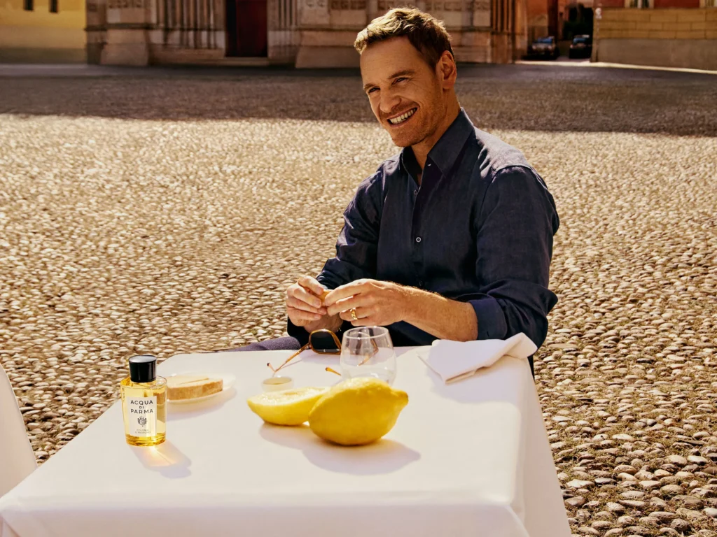 A man in a dark shirt sits at an outdoor table with a large lemon, a drink, and a bottle of Acqua di Parma.