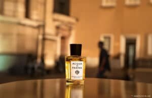 A bottle of Acqua di Parma Colonia perfume on a reflective surface, with blurred outdoor urban background.