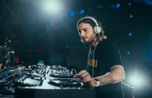 A DJ wearing headphones is focused while mixing music at a DJ booth, illuminated by stage lights.