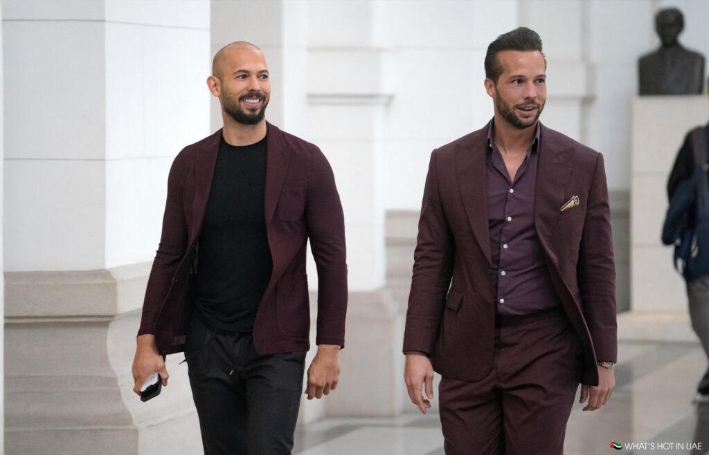 Two men in matching burgundy suits walk indoors, smiling and holding items.