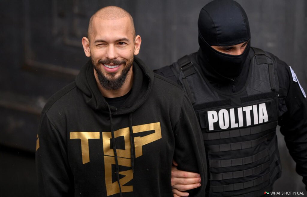 Andrew Tate winking and smiling is escorted by a masked officer wearing a "Politia" vest.