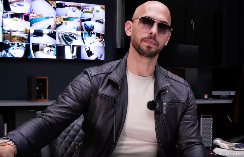 Andrew Tate wearing sunglasses and a leather jacket sits in front of a screen displaying multiple security camera feeds.