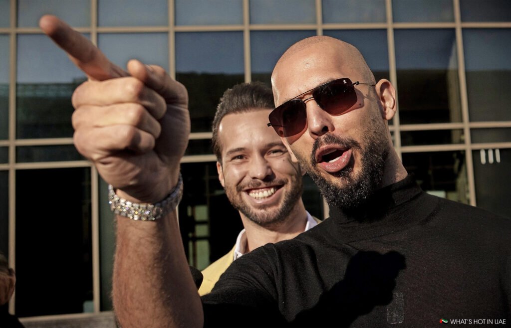 Two men, one wearing sunglasses, with a modern building in the background; the front man points forward animatedly.