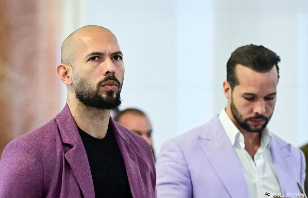 Andrew and Tristan Tate in formal attire, one wearing a pink suit and another in a light purple jacket, standing indoors.