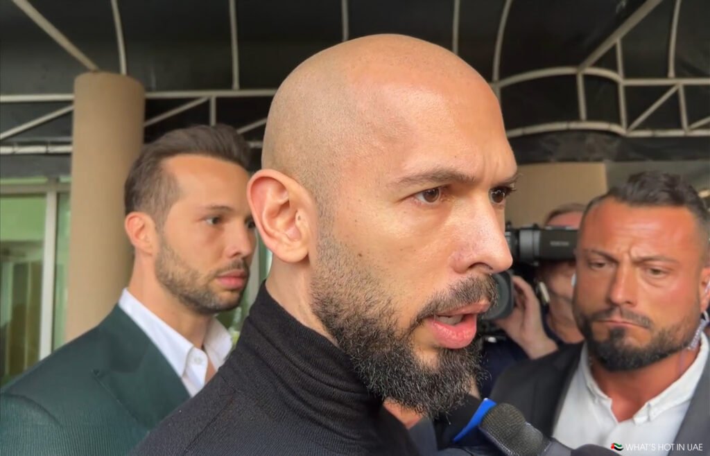 A man with a bald head and beard speaks into a microphone surrounded by reporters and cameras.