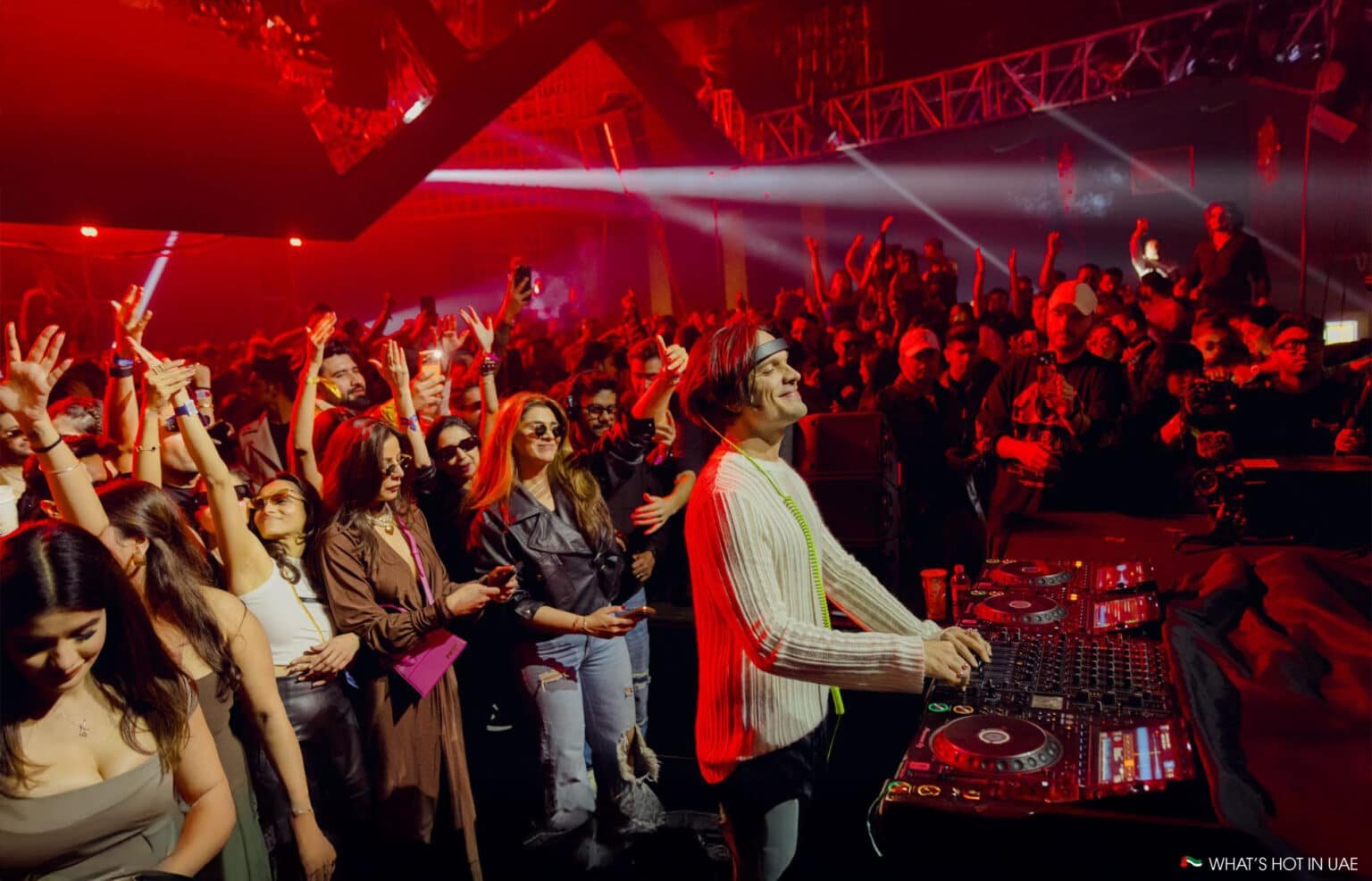 A DJ performing on stage with a lively crowd dancing under red lights.