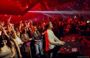 A DJ performing on stage with a lively crowd dancing under red lights.