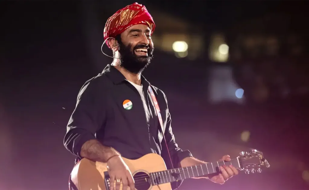Arijit Singh wearing a red turban and a shirt with an Indian flag pin plays an acoustic guitar while smiling.