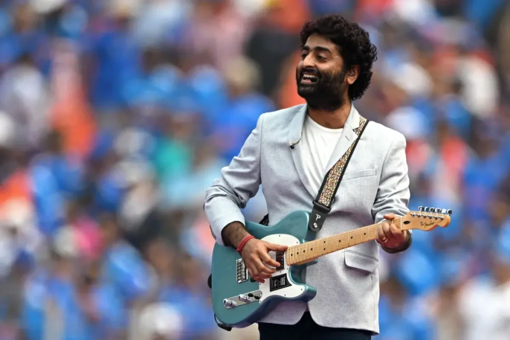 Arijit Singh playing a light blue electric guitar outdoors, dressed in a gray blazer, with a blurred crowd in the background.