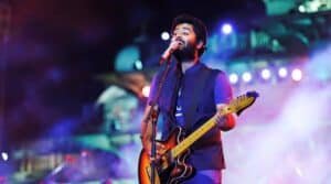 A man singing and playing an electric guitar on stage with colorful lights and smoke effects in the background.