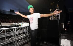 A DJ on stage with outstretched arms, wearing a cap and a graphic t-shirt, before a large crowd.