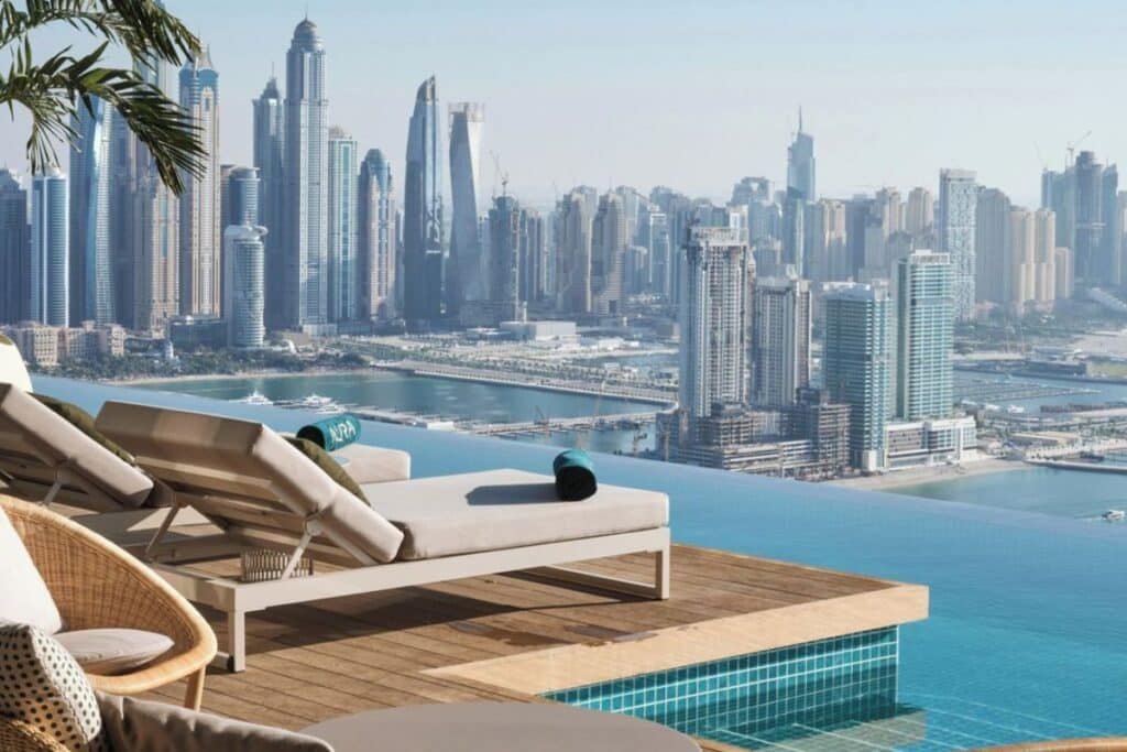 Two lounge chairs with towels overlook a blue infinity pool and a city skyline with tall skyscrapers in the background.