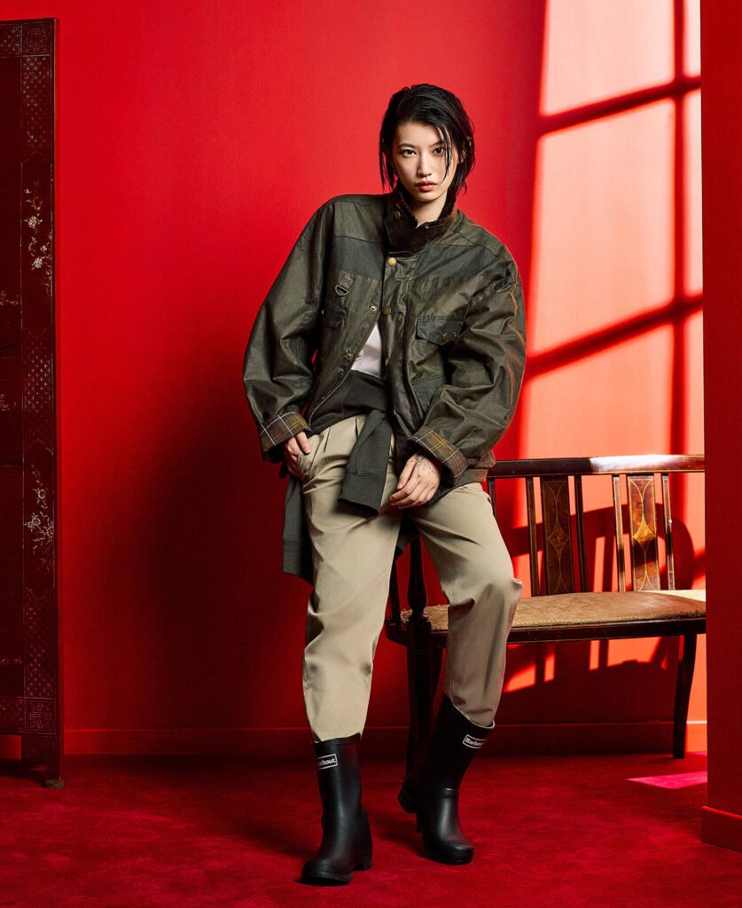 A person in a rugged jacket and boots poses indoors against a red wall, with a decorative screen and chair nearby.