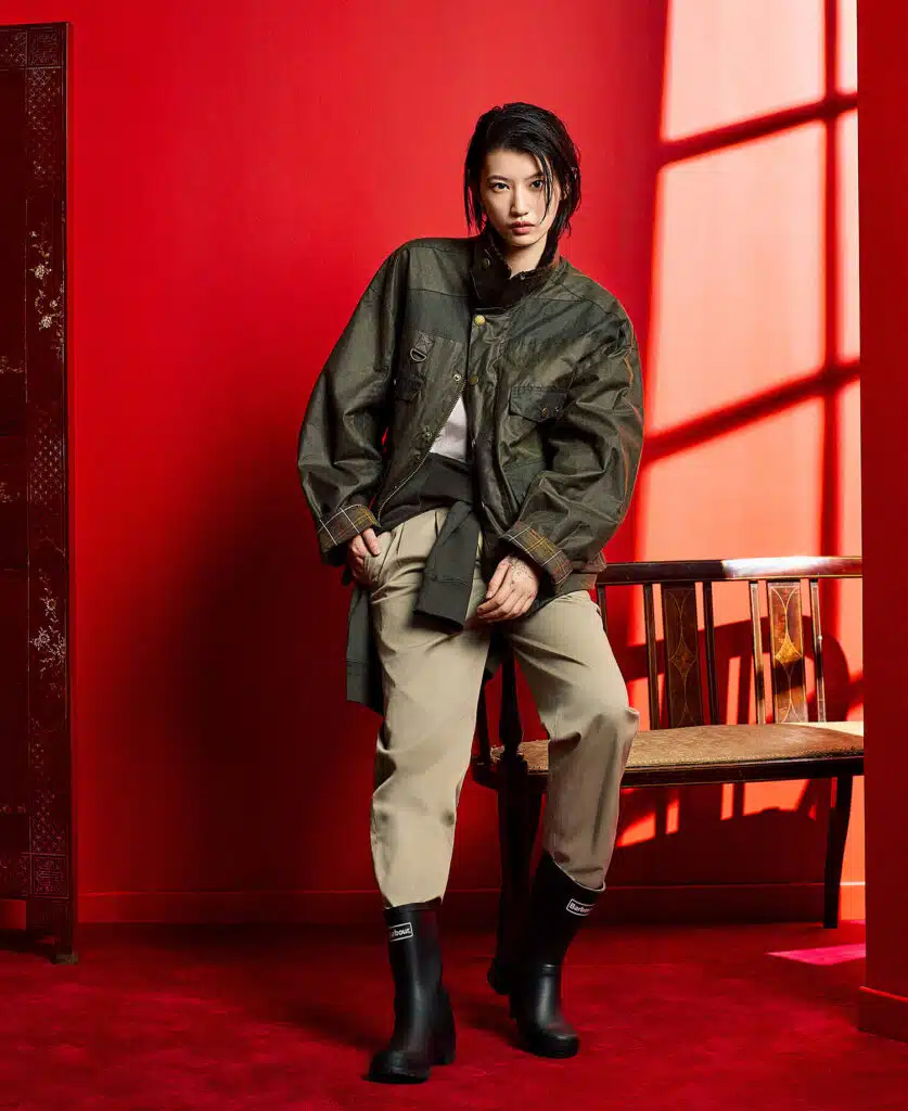 A person in a rugged jacket and boots poses indoors against a red wall, with a decorative screen and chair nearby.