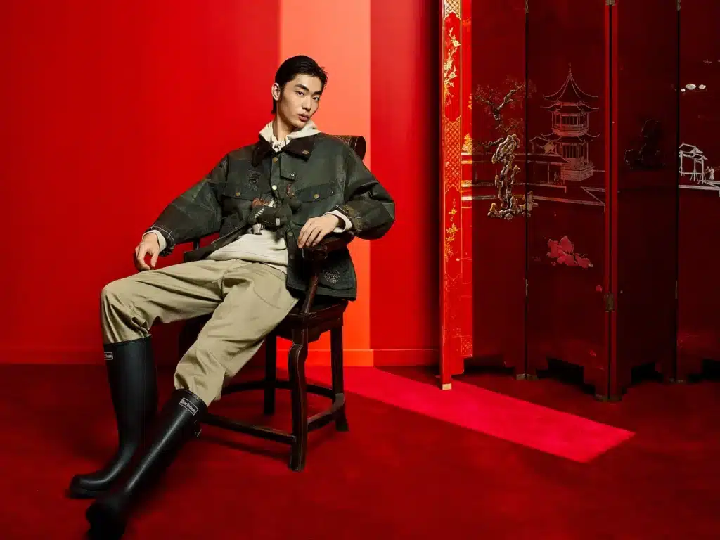 Person sitting in a chair against a red backdrop, wearing a Barbour x Feng Chen Wang green jacket, beige pants, and black boots.