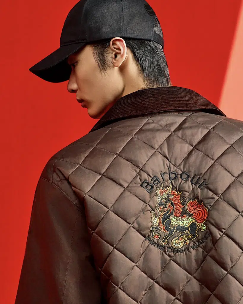 A person wearing a quilted Barbour x Feng Chen Wang brown jacket with a dragon embroidered on the back, set against a lively red background.