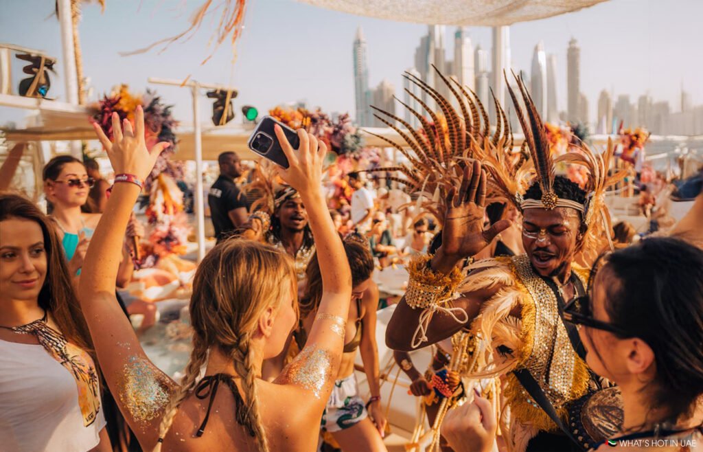 People in vibrant, feathered costumes dance energetically at a lively outdoor event with a city skyline in the background.
