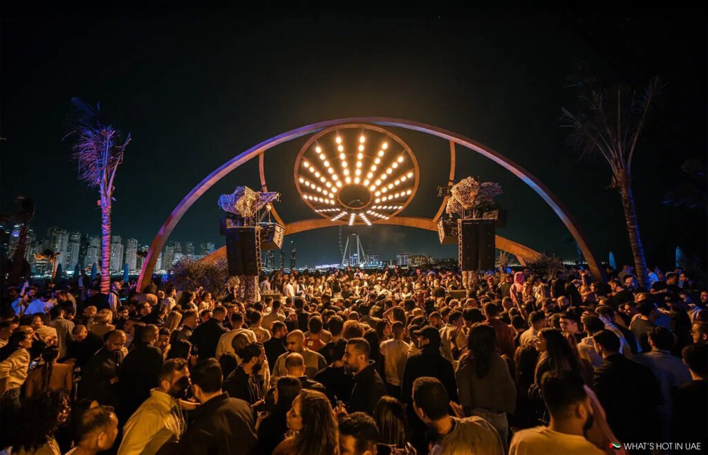 A large crowd gathers at an outdoor nightlife event under a circular illuminated structure at night.