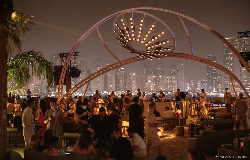A bustling outdoor Be Beach DXB with people socializing under a large decorative arch, city skyline illuminated in the background.