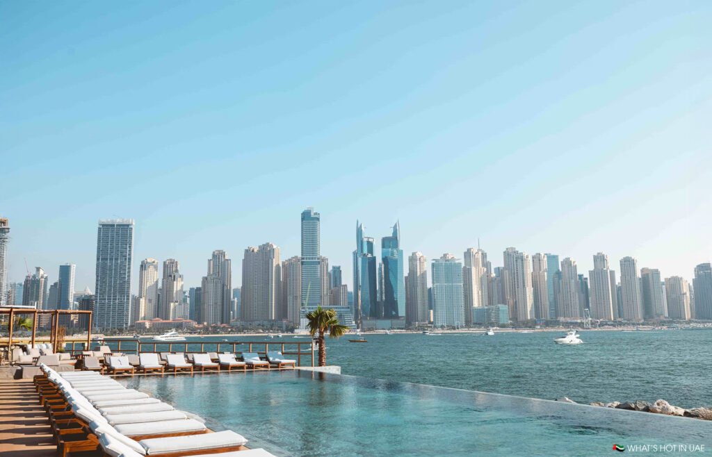 Infinity pool at Be Beach DXB overlooking a vast water body with a city skyline in the background under a clear blue sky.