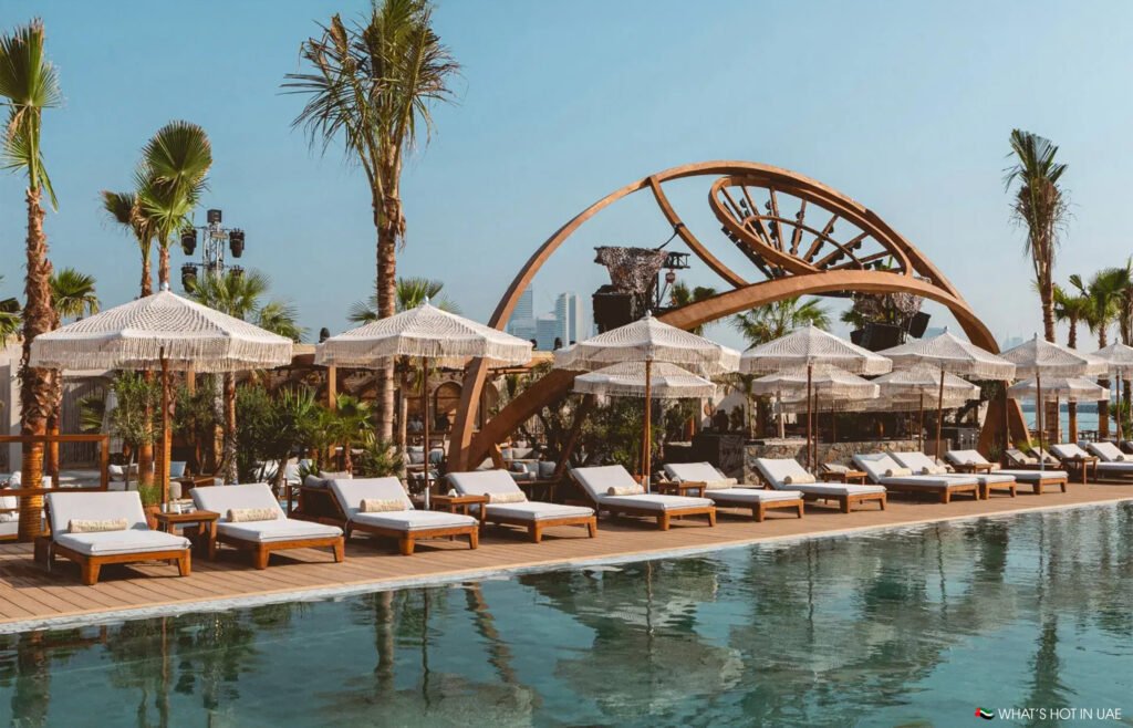 Poolside area at Be Beach DXB with white lounge chairs, umbrellas, and palm trees, featuring a decorative wooden arch structure.