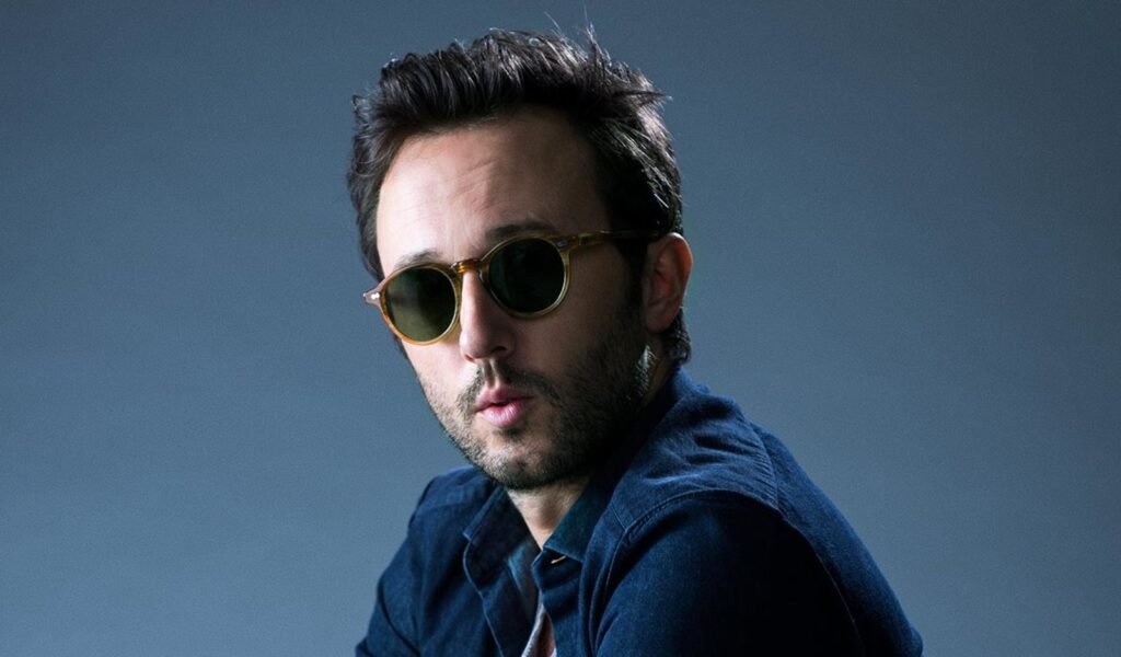 A man with short dark hair and a beard wearing round sunglasses and a blue denim shirt against a neutral background.