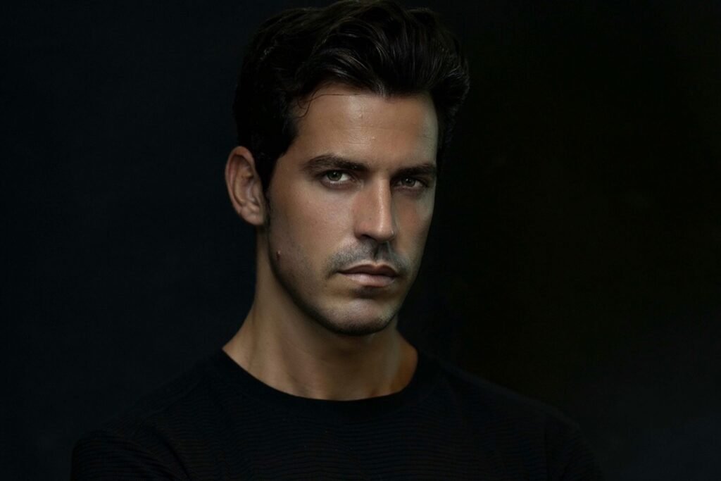 A man with dark hair and a serious expression, wearing a black shirt against a dark background.