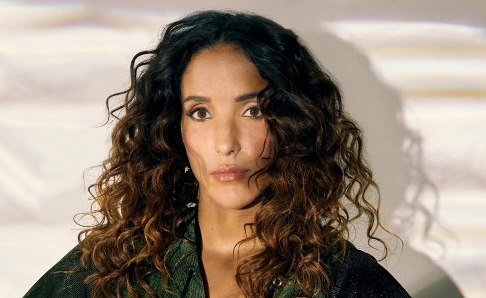 A person with long, curly hair wearing a dark top, looking directly at the camera with a neutral expression.