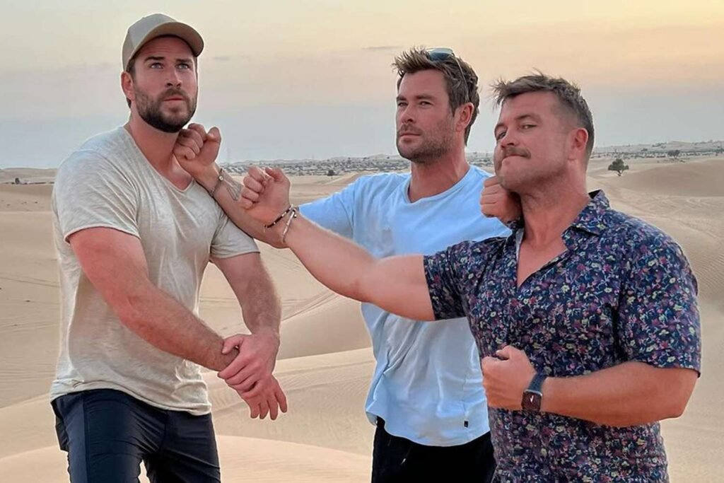 Three men jokingly pose in a desert with one pretending to choke another as a playful gesture.