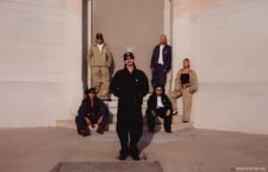 A group of six people poses in front of a building, each wearing casual streetwear.