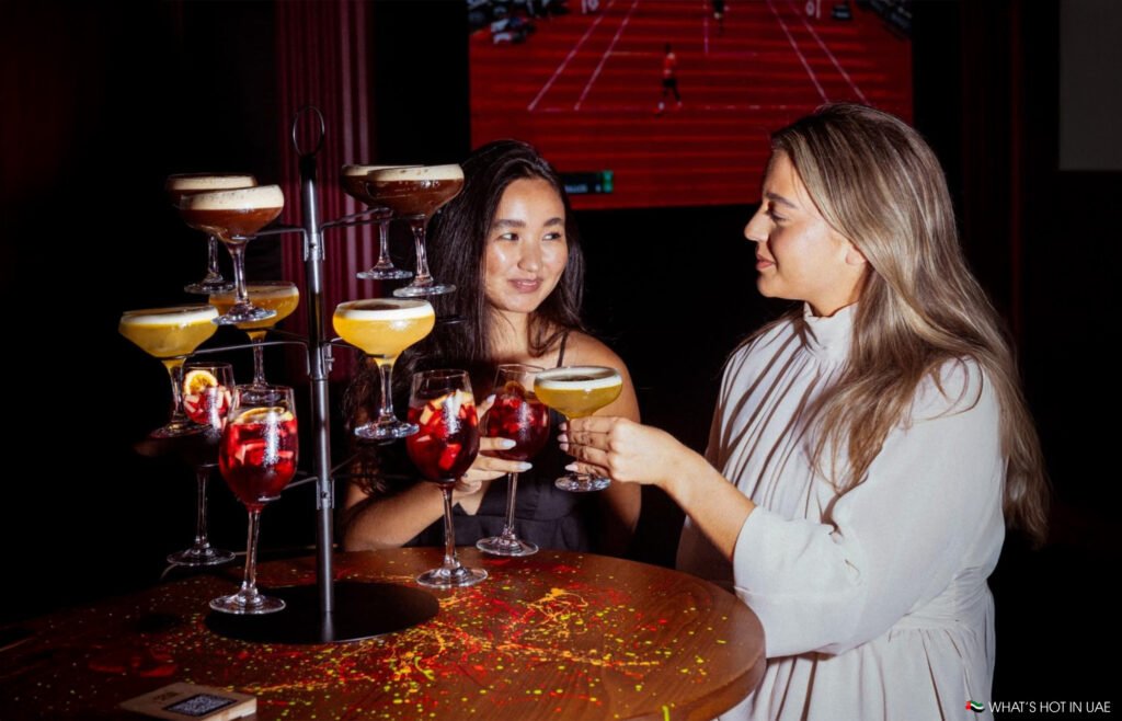 Two women clinking glasses surrounded by a Best Happy Hours tiered display of various colorful cocktails.