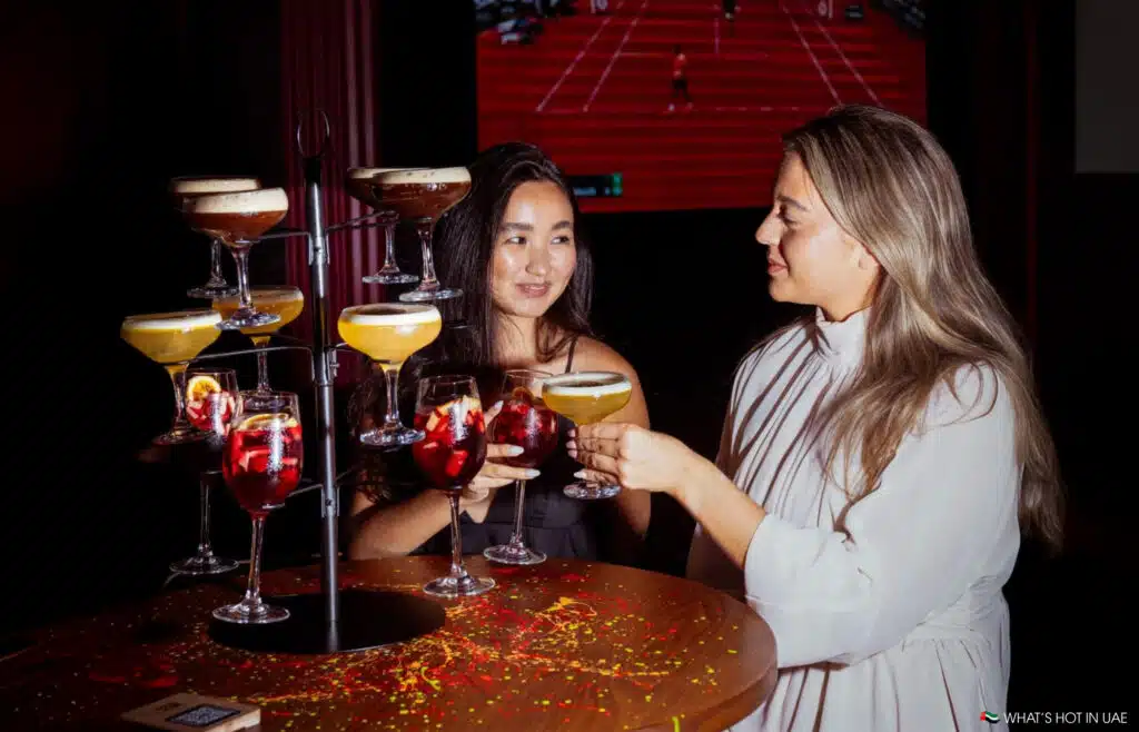 Two women clinking glasses surrounded by a Best Happy Hours tiered display of various colorful cocktails.