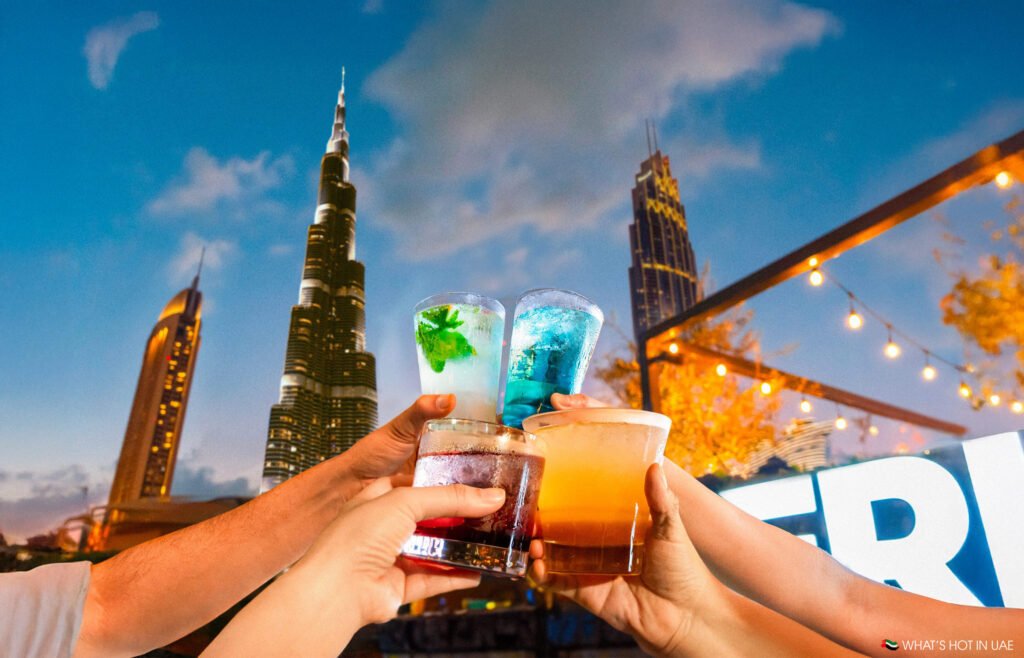 Four hands holding drinks raised in a toast, against a backdrop of tall city buildings and decorative lights.